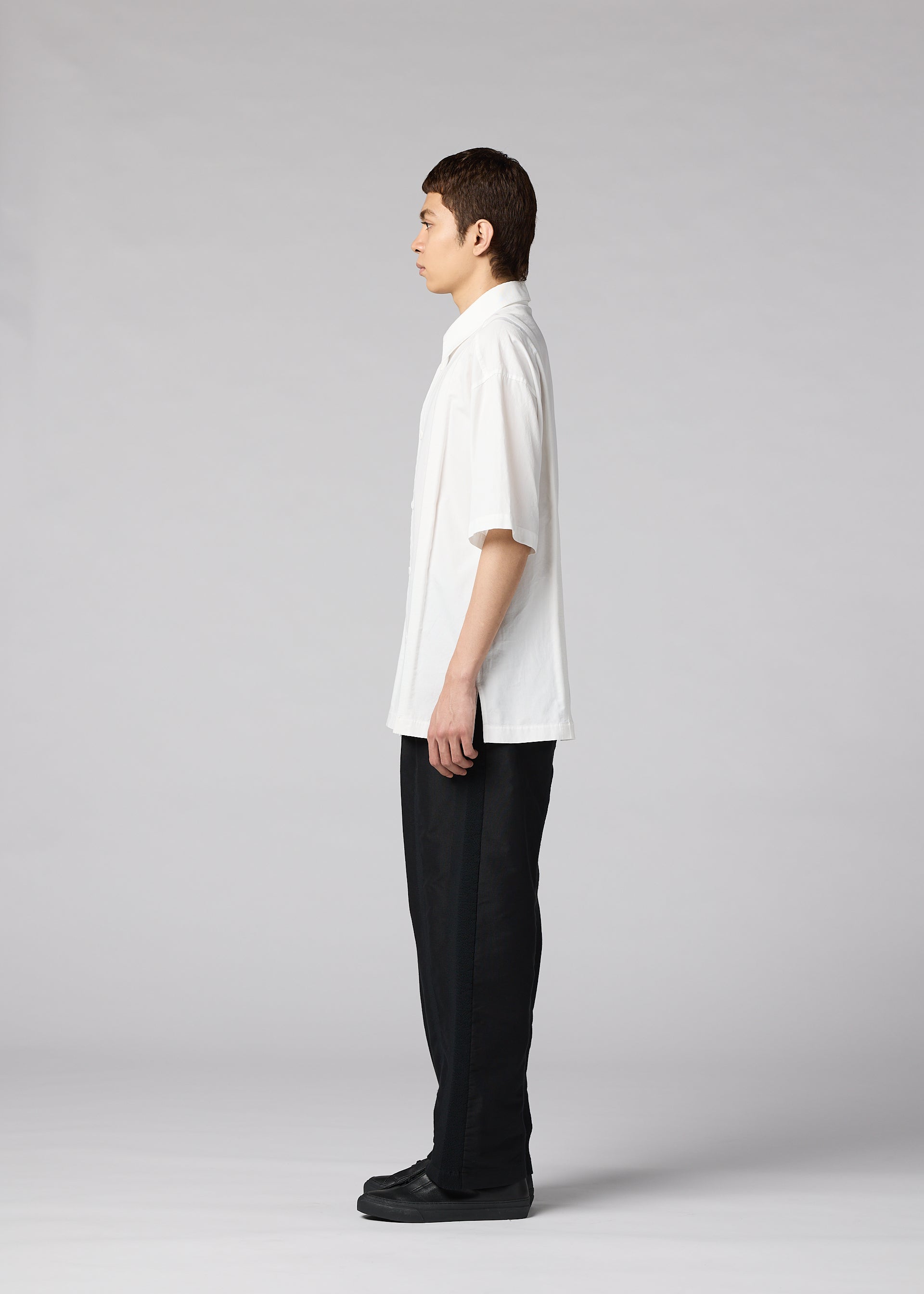 Zoomed view A person with short dark hair stands in profile, facing left, wearing the IM MEN CLAY SHIRT (loose white cotton, short sleeves), black pants, and black shoes against a plain light gray background.