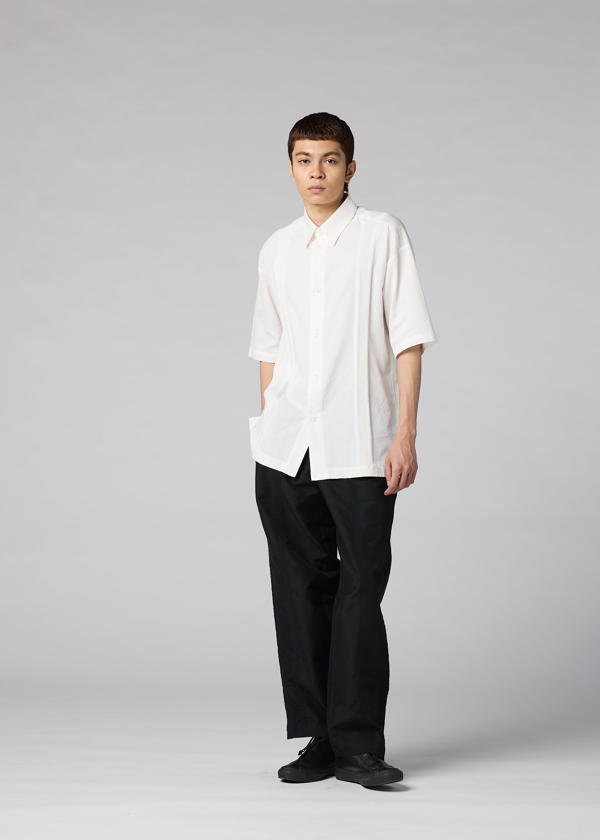 Zoomed view A person stands against a plain gray background, wearing the IM MEN CLAY SHIRT— a loose-fitting white cotton short-sleeve shirt—paired with black pants and shoes, one hand in their pocket and a neutral expression.