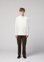 A person with short, curly hair faces forward against a plain light grey background, wearing the IM MEN 140/2 SHIRT—a white, long-sleeved cotton shirt—paired with brown trousers and black shoes.