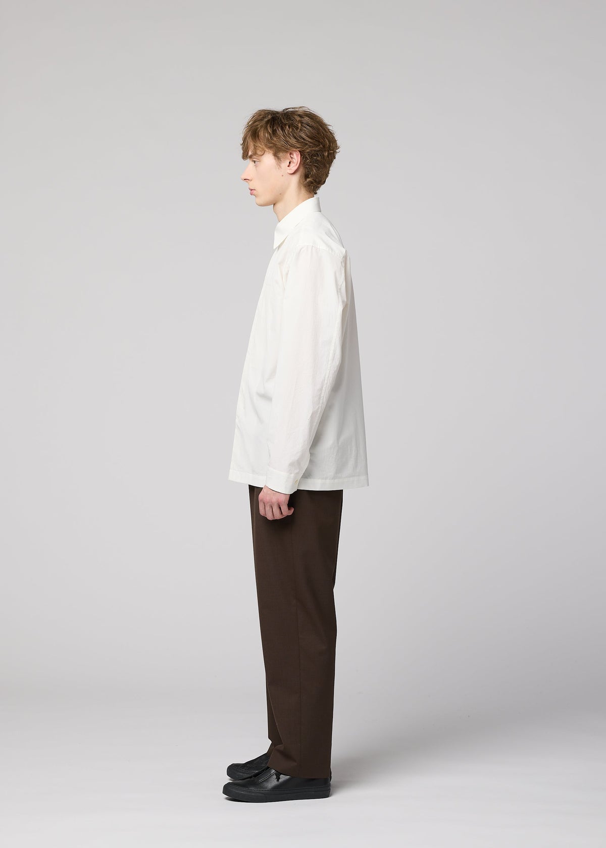 A young man stands in profile against a light gray background, wearing the IM MEN 140/2 SHIRT—a crisp white long-sleeved shirt made from fine cotton—paired with dark brown pants and black shoes. view 7 of 9