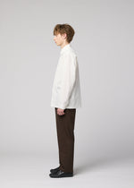 A young man stands in profile against a light gray background, wearing the IM MEN 140/2 SHIRT—a crisp white long-sleeved shirt made from fine cotton—paired with dark brown pants and black shoes.