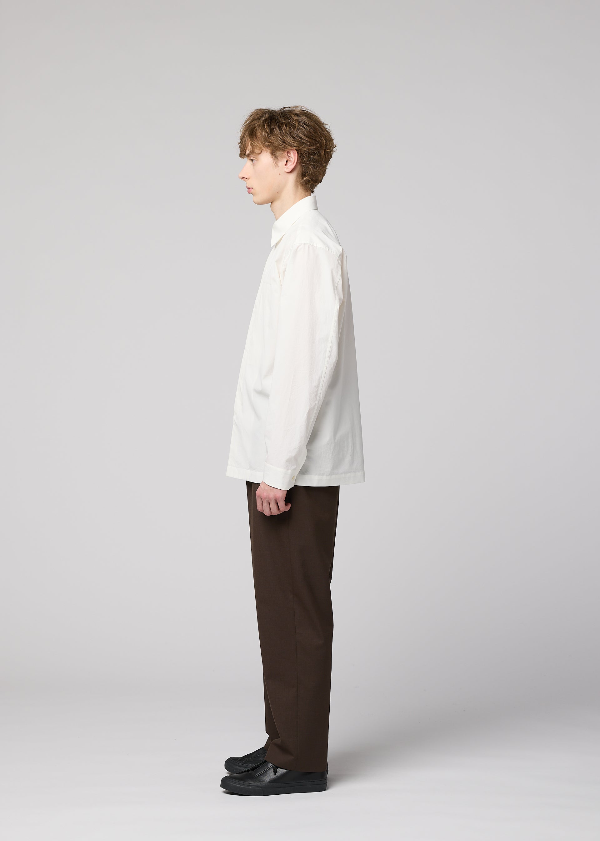Zoomed view A young man stands in profile against a light gray background, wearing the IM MEN 140/2 SHIRT—a crisp white long-sleeved shirt made from fine cotton—paired with dark brown pants and black shoes.
