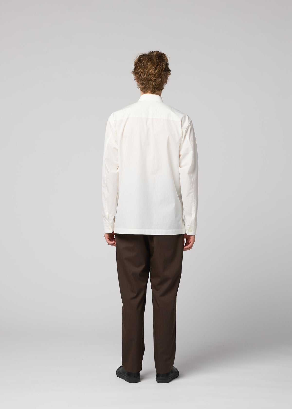 A person with curly hair faces away from the camera, wearing the IM MEN 140/2 SHIRT in white, brown pants, and black shoes against a plain light gray background. view 8 of 9