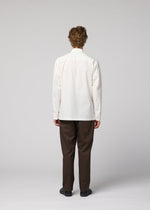 A person with curly hair faces away from the camera, wearing the IM MEN 140/2 SHIRT in white, brown pants, and black shoes against a plain light gray background.