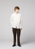 A person with curly hair stands against a plain background wearing the IM MEN 140/2 SHIRT, paired with brown pants and black shoes. One hand is in their pocket as they look to the side.