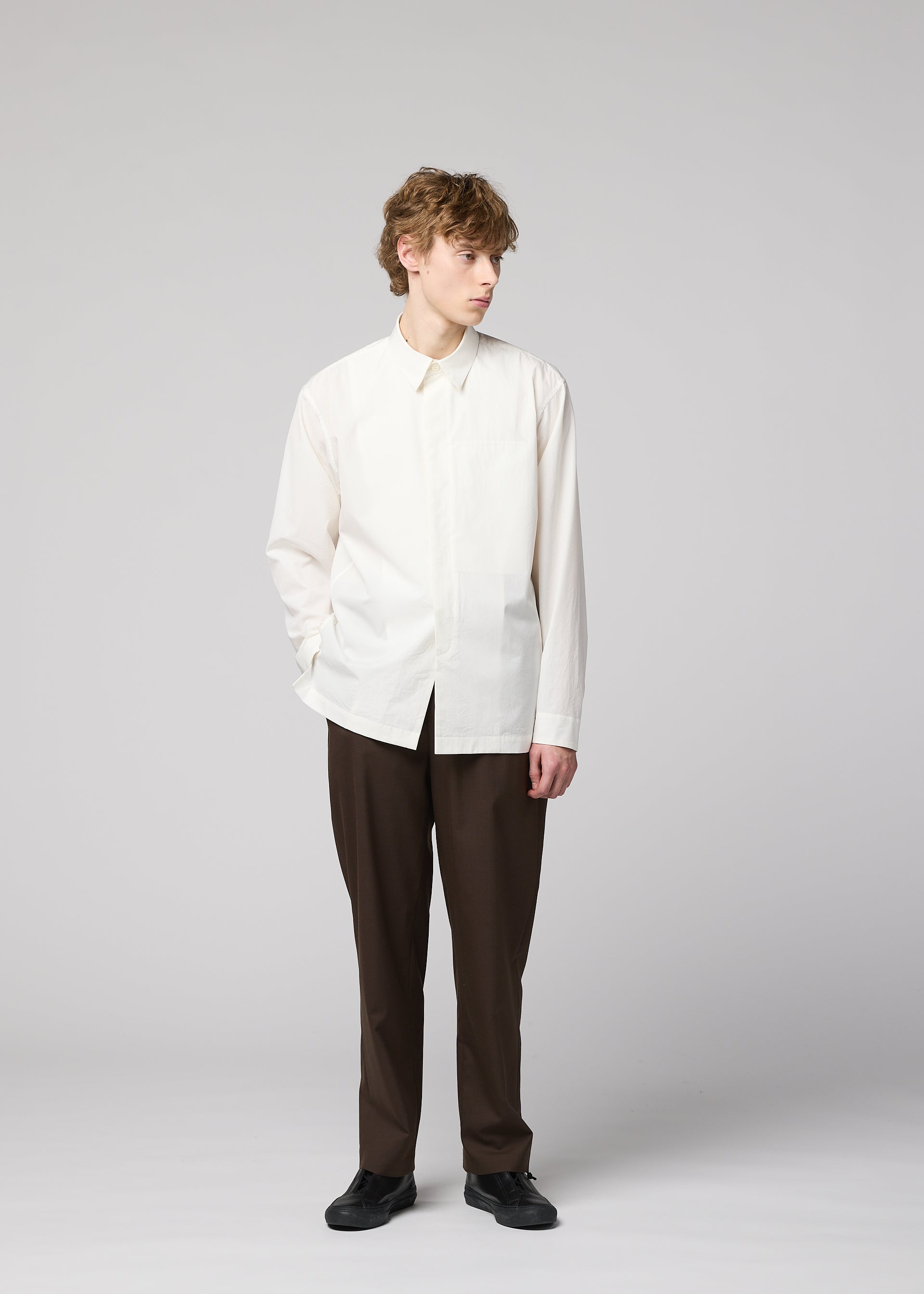 Zoomed view A person with curly hair stands against a plain background wearing the IM MEN 140/2 SHIRT, paired with brown pants and black shoes. One hand is in their pocket as they look to the side.