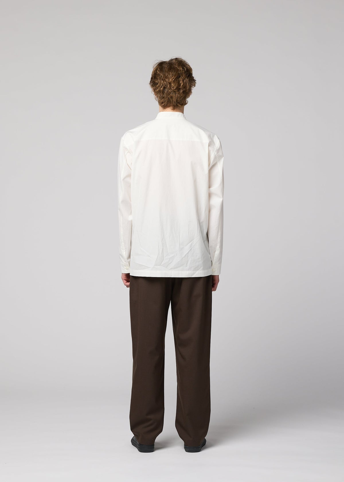 A person with short, curly hair stands facing away from the camera against a plain light gray background, wearing the IM MEN 140/2 SHIRT in white with long sleeves and stand-up collar, paired with loose-fitting brown pants. view 8 of 9