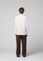 A person with short, curly hair stands facing away from the camera against a plain light gray background, wearing the IM MEN 140/2 SHIRT in white with long sleeves and stand-up collar, paired with loose-fitting brown pants.