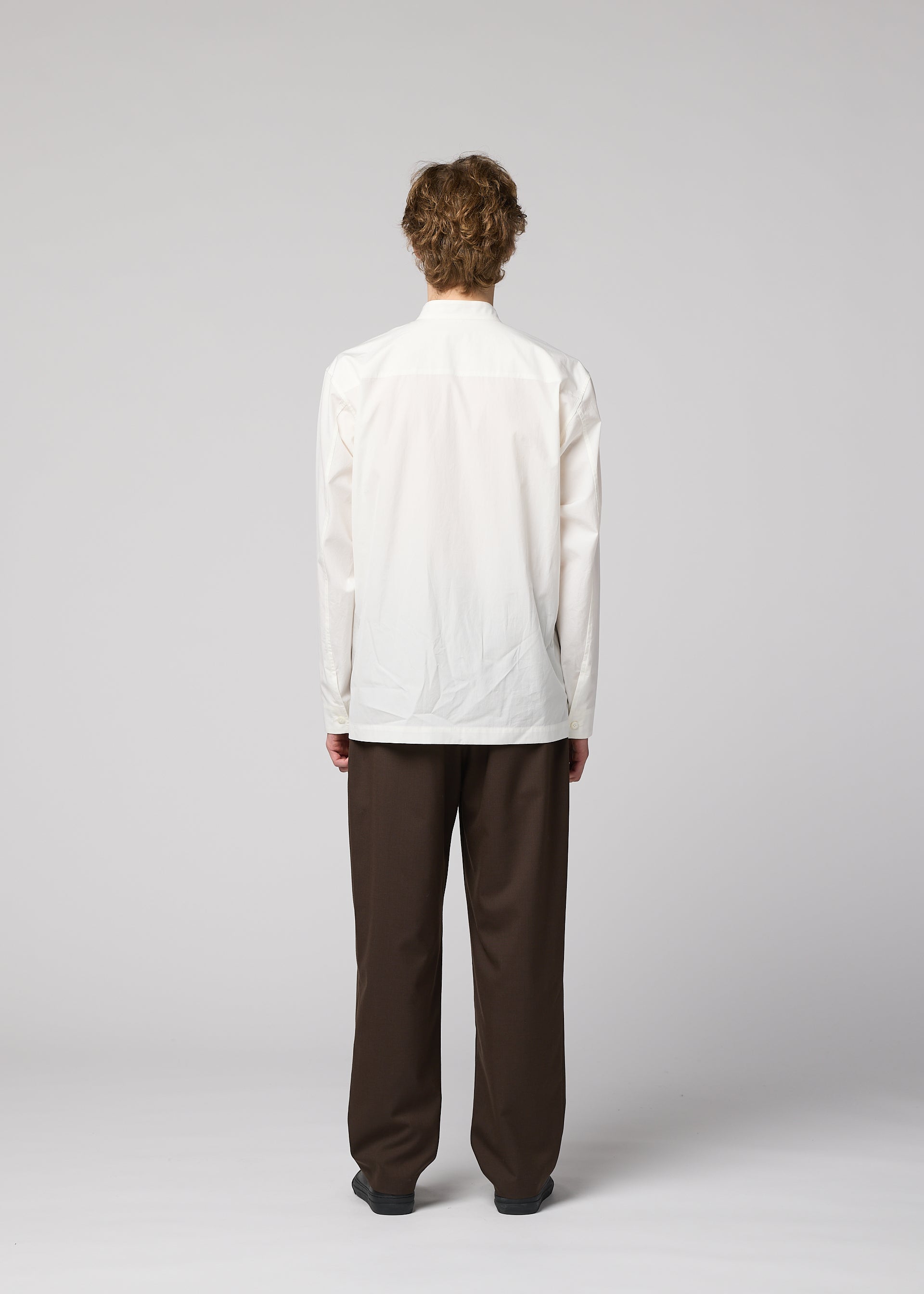 Zoomed view A person with short, curly hair stands facing away from the camera against a plain light gray background, wearing the IM MEN 140/2 SHIRT in white with long sleeves and stand-up collar, paired with loose-fitting brown pants.