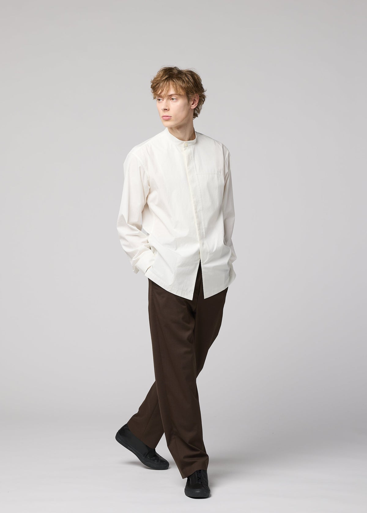 A person with light brown hair stands against a plain light background, wearing the IM MEN 140/2 SHIRT, loose brown trousers, and black shoes, with hands in pockets and looking to the side. view 9 of 9