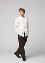 A person with light brown hair stands against a plain light background, wearing the IM MEN 140/2 SHIRT, loose brown trousers, and black shoes, with hands in pockets and looking to the side.