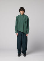 A person with medium-length dark hair stands against a plain light gray background, wearing the IM MEN 140/2 SHIRT in green, paired with dark teal wide-leg pants and black shoes.