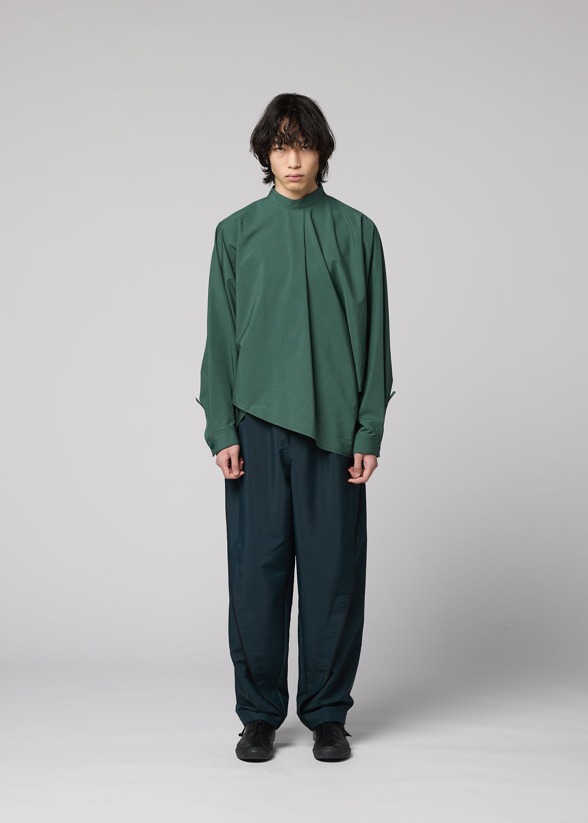 Zoomed view A person with medium-length dark hair stands against a plain light gray background, wearing the IM MEN 140/2 SHIRT in green, paired with dark teal wide-leg pants and black shoes.