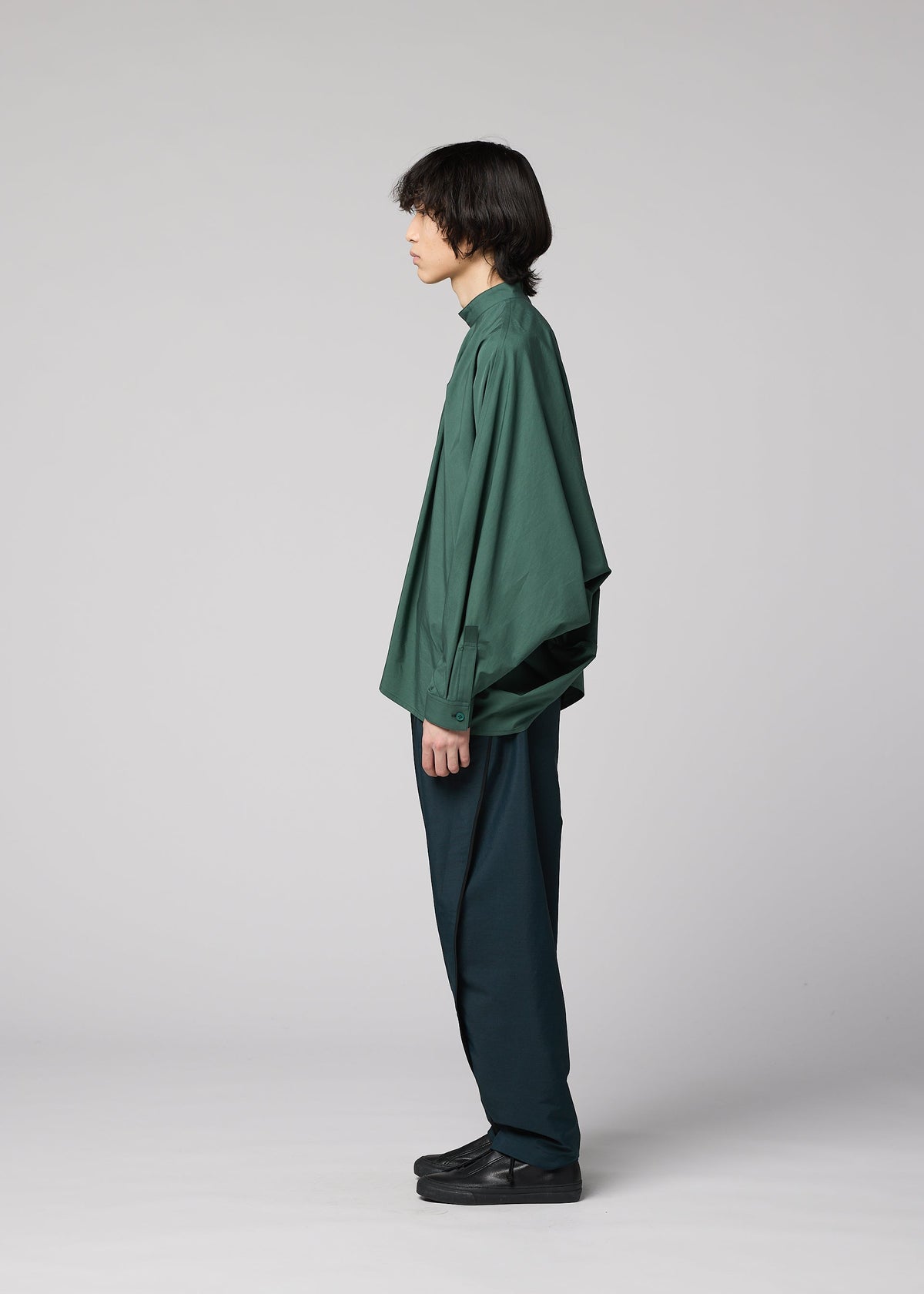 A person with medium-length dark hair stands in profile against a plain light gray background, wearing the IM MEN 140/2 SHIRT in green, paired with dark green pants and black shoes. view 7 of 9