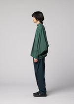 A person with medium-length dark hair stands in profile against a plain light gray background, wearing the IM MEN 140/2 SHIRT in green, paired with dark green pants and black shoes.