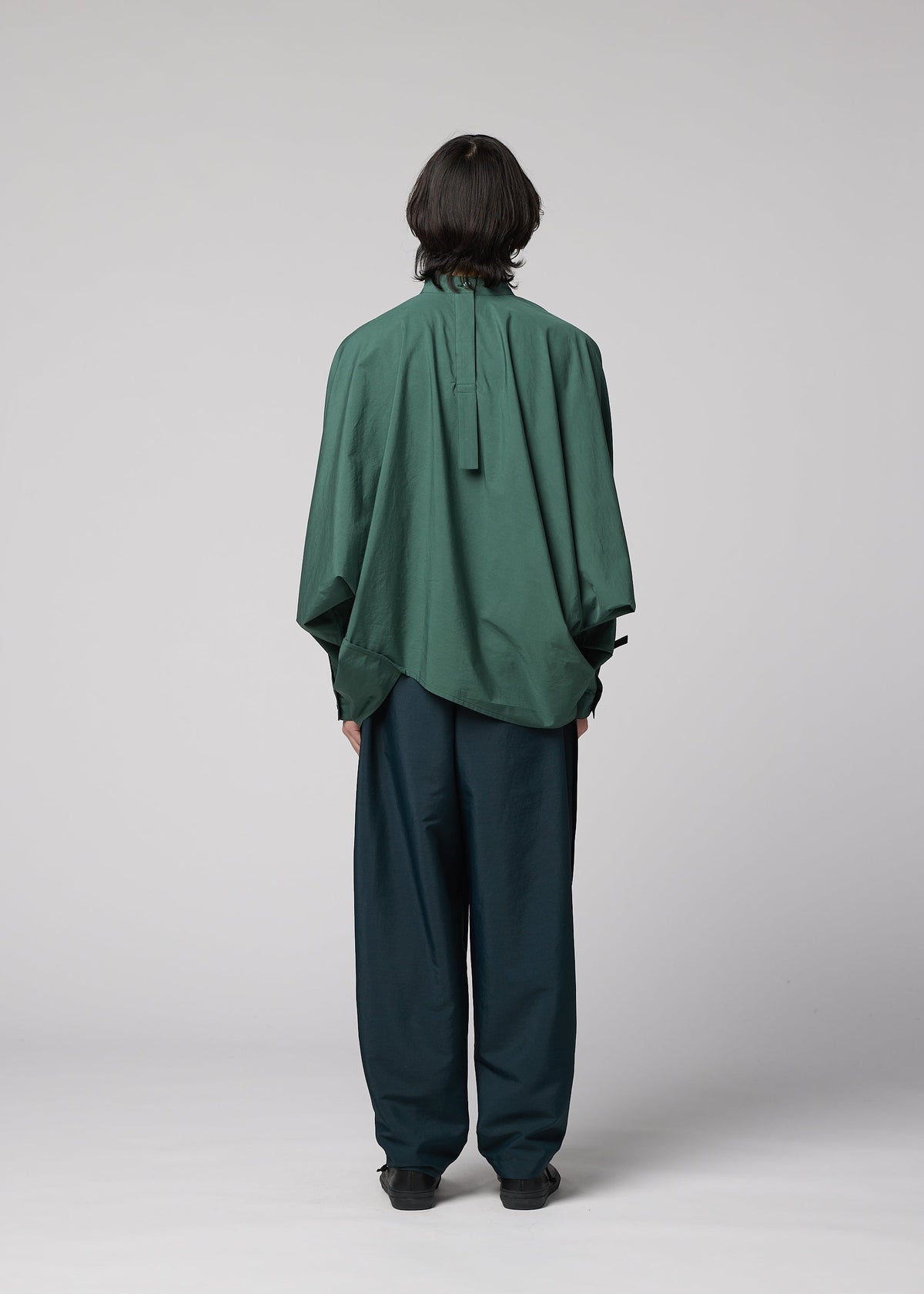 A person with shoulder-length dark hair faces away, wearing the IM MEN 140/2 SHIRT—an oversized green band collar shirt with a relaxed fit—and loose dark green trousers against a plain light gray background. view 8 of 9