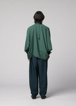 A person with shoulder-length dark hair faces away, wearing the IM MEN 140/2 SHIRT—an oversized green band collar shirt with a relaxed fit—and loose dark green trousers against a plain light gray background.