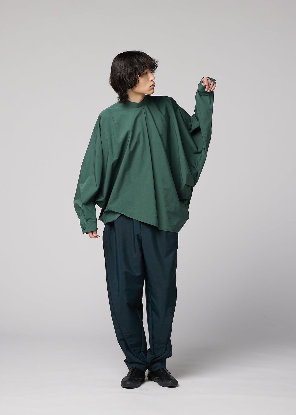 A person with medium-length dark hair wears the IM MEN 140/2 SHIRT, styled oversized with a band collar and relaxed silhouette, paired with loose dark pants, standing against a plain gray background while looking to the side. view 9 of 9