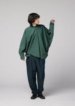A person with medium-length dark hair wears the IM MEN 140/2 SHIRT, styled oversized with a band collar and relaxed silhouette, paired with loose dark pants, standing against a plain gray background while looking to the side.