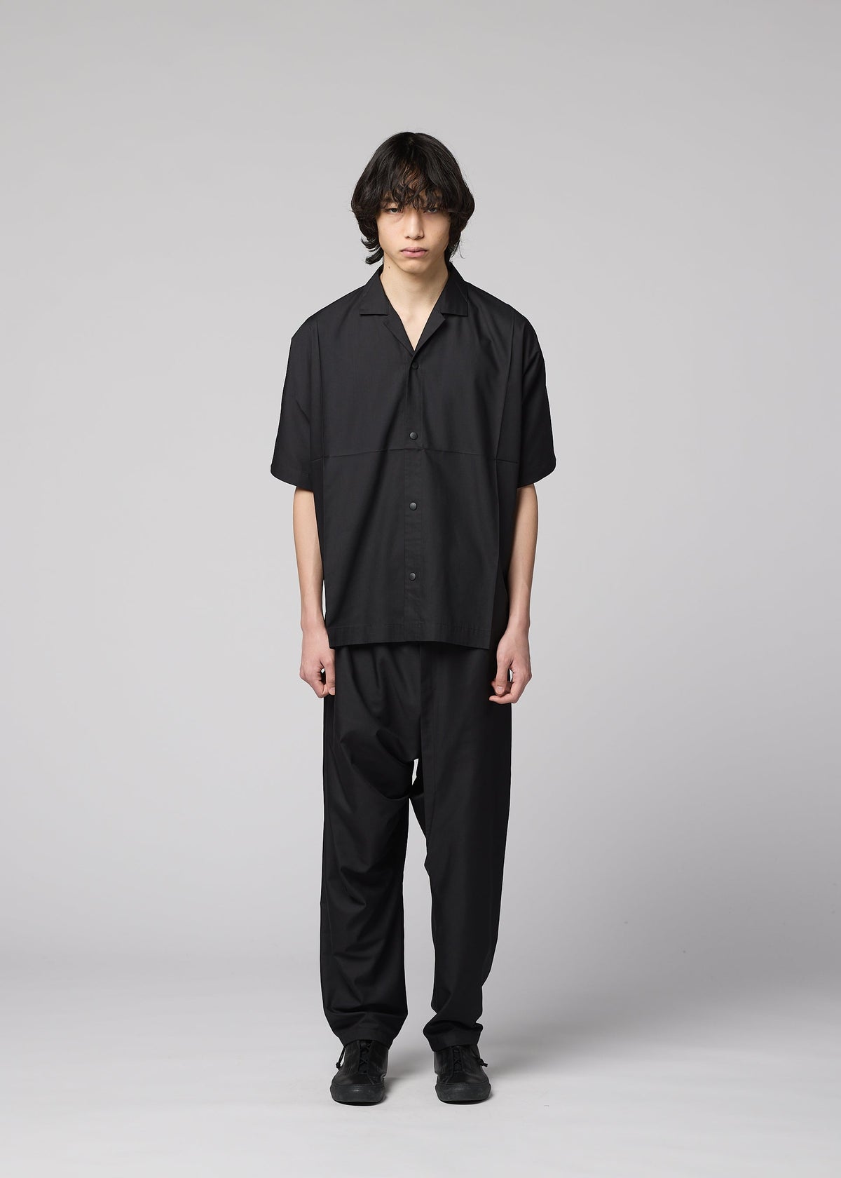 A person with medium-length dark hair faces forward, wearing the IM MEN COMPACT SHIRT in black, made of light wrinkle-resistant material, along with matching black pants and shoes, against a plain light gray background. view 7 of 10