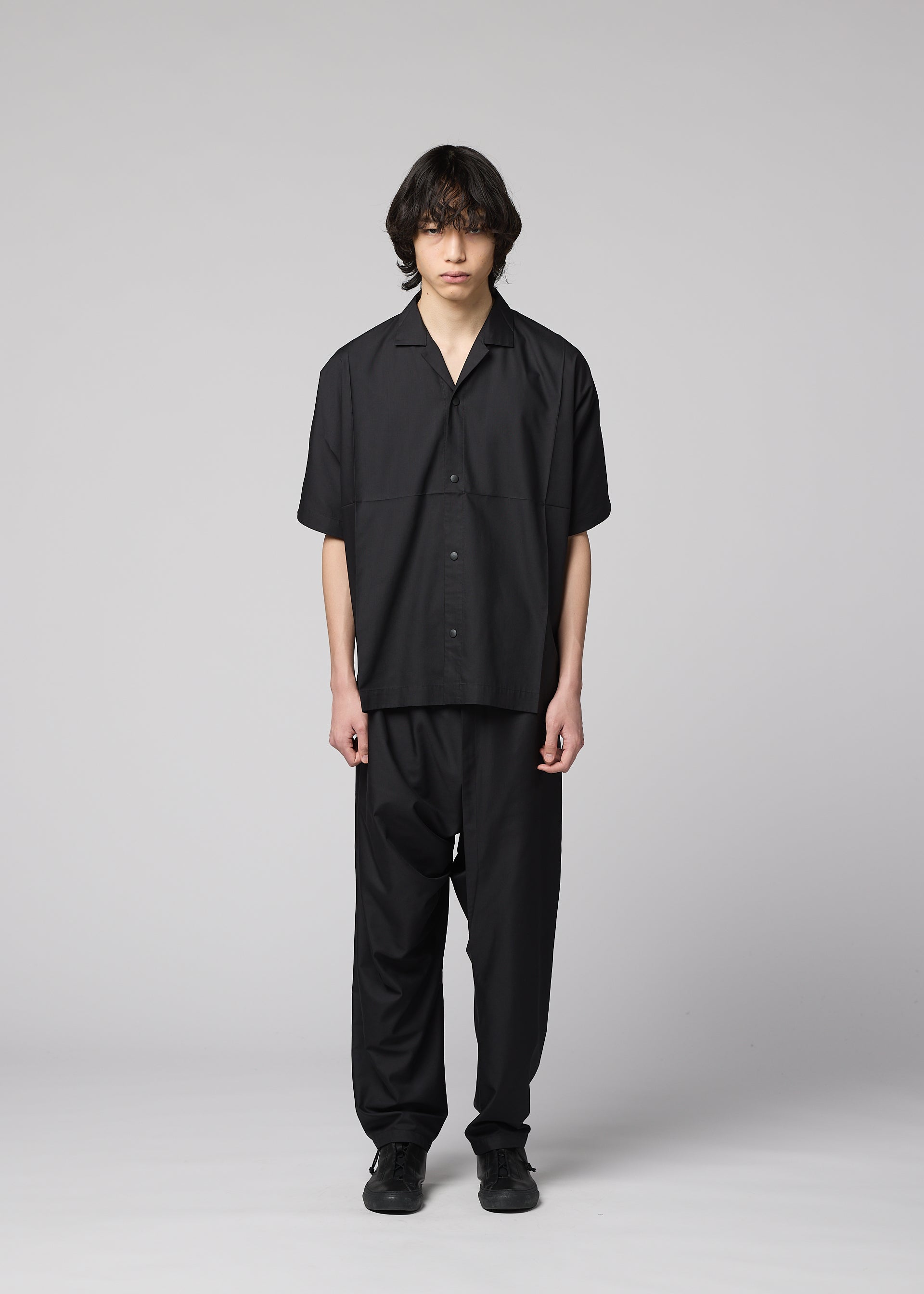 Zoomed view A person with medium-length dark hair faces forward, wearing the IM MEN COMPACT SHIRT in black, made of light wrinkle-resistant material, along with matching black pants and shoes, against a plain light gray background.