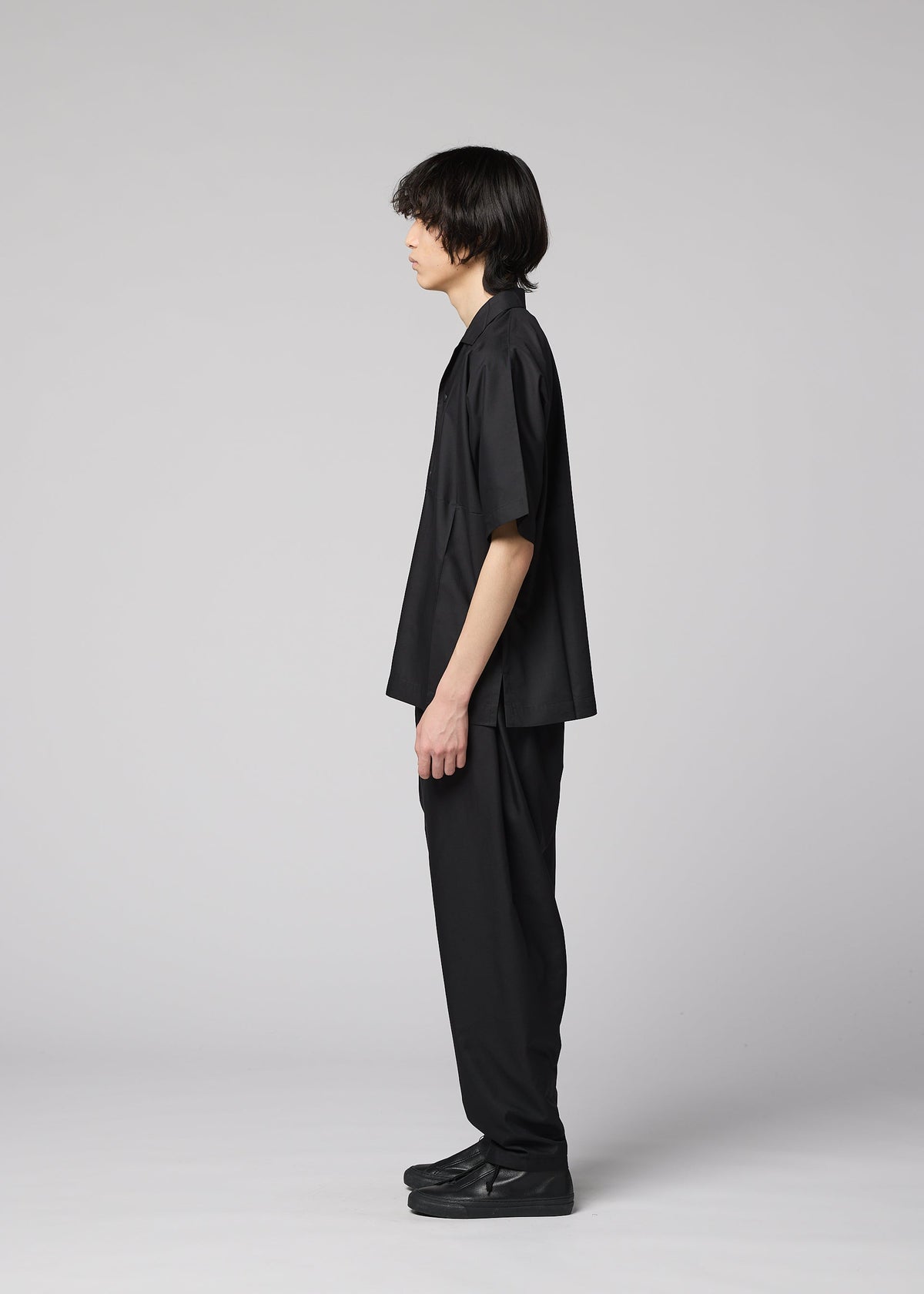 A person with medium-length dark hair stands in profile facing left, wearing the IM MEN COMPACT SHIRT—wrinkle-resistant and loose-fitting—along with black pants and shoes, against a plain light gray background. view 8 of 10