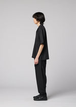 A person with medium-length dark hair stands in profile facing left, wearing the IM MEN COMPACT SHIRT—wrinkle-resistant and loose-fitting—along with black pants and shoes, against a plain light gray background.