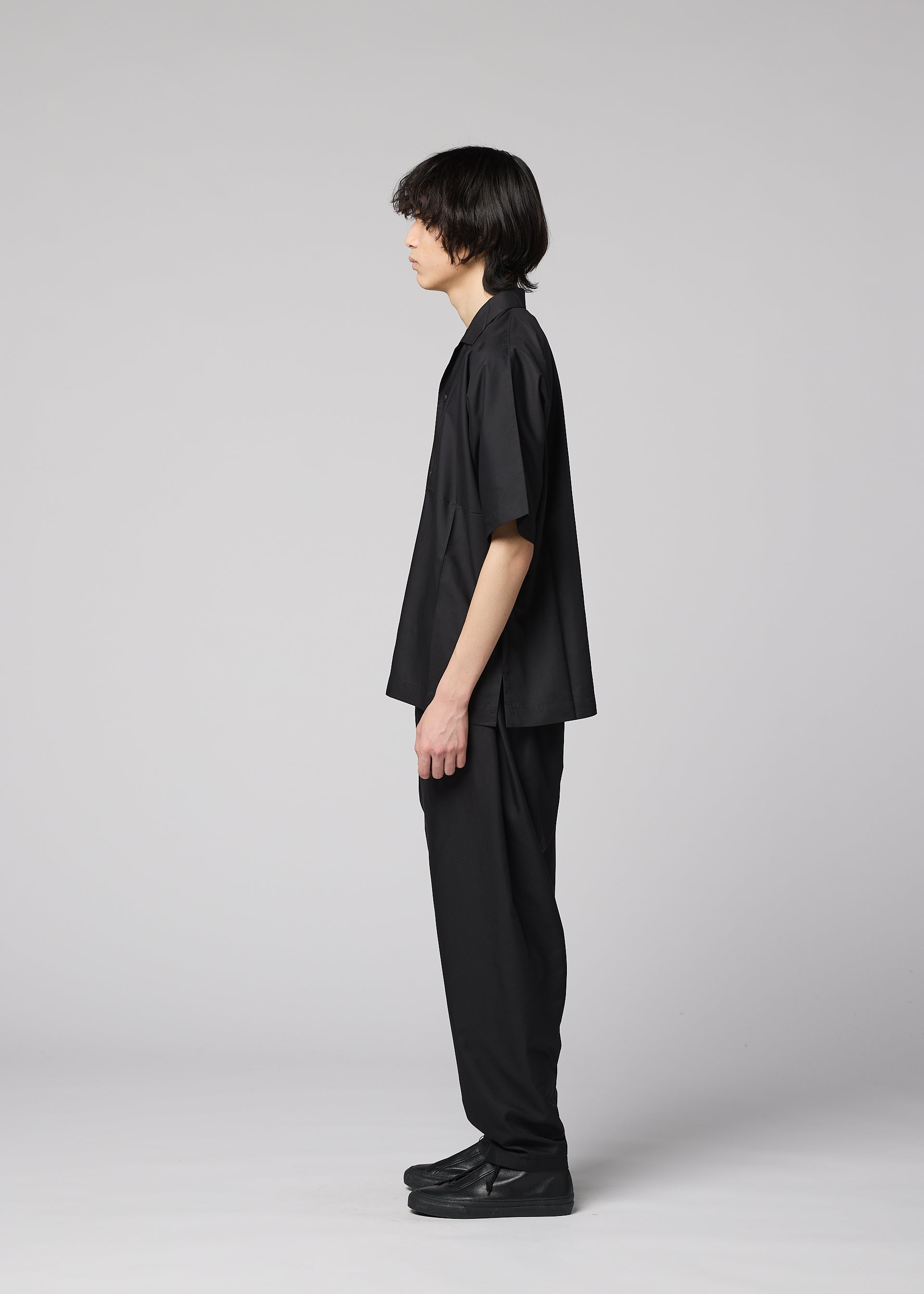 Zoomed view A person with medium-length dark hair stands in profile facing left, wearing the IM MEN COMPACT SHIRT—wrinkle-resistant and loose-fitting—along with black pants and shoes, against a plain light gray background.