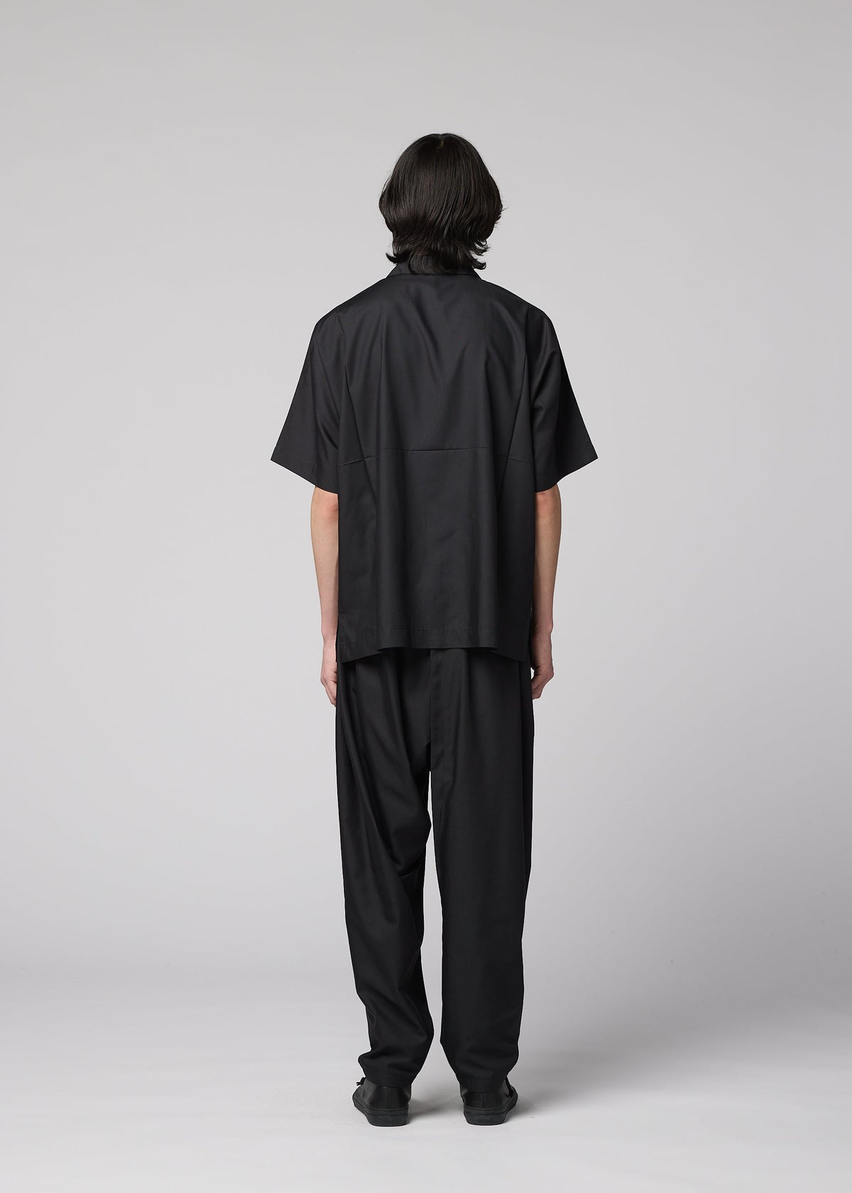 A person with straight, dark hair faces away from the camera, wearing the IM MEN COMPACT SHIRT—a wrinkle-resistant, lightweight black short-sleeved shirt—paired with matching black pants, set against a plain gray background. view 9 of 10