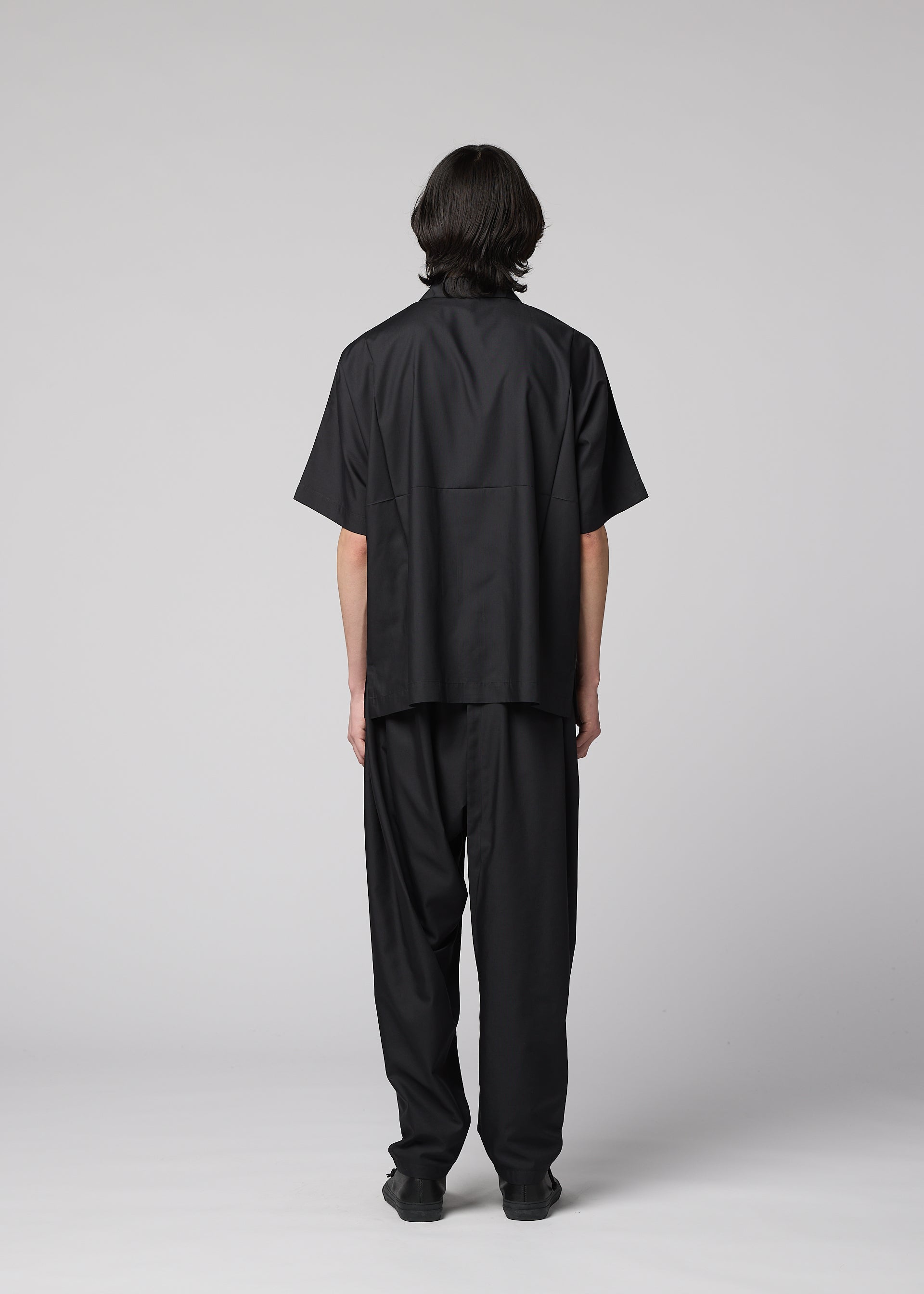 Zoomed view A person with straight, dark hair faces away from the camera, wearing the IM MEN COMPACT SHIRT—a wrinkle-resistant, lightweight black short-sleeved shirt—paired with matching black pants, set against a plain gray background.