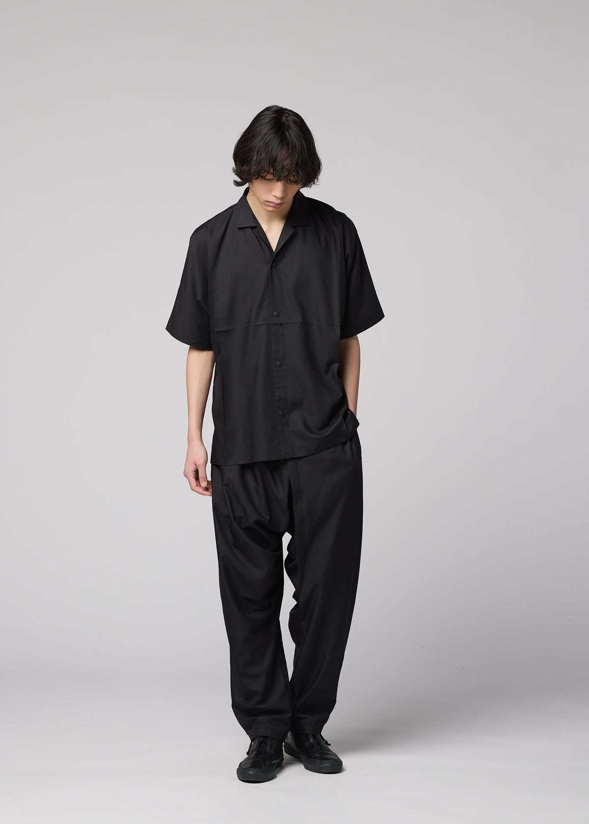 A person with medium-length dark hair stands looking down, wearing the IM MEN COMPACT SHIRT and matching black pants with black shoes, against a plain light gray background. view 10 of 10