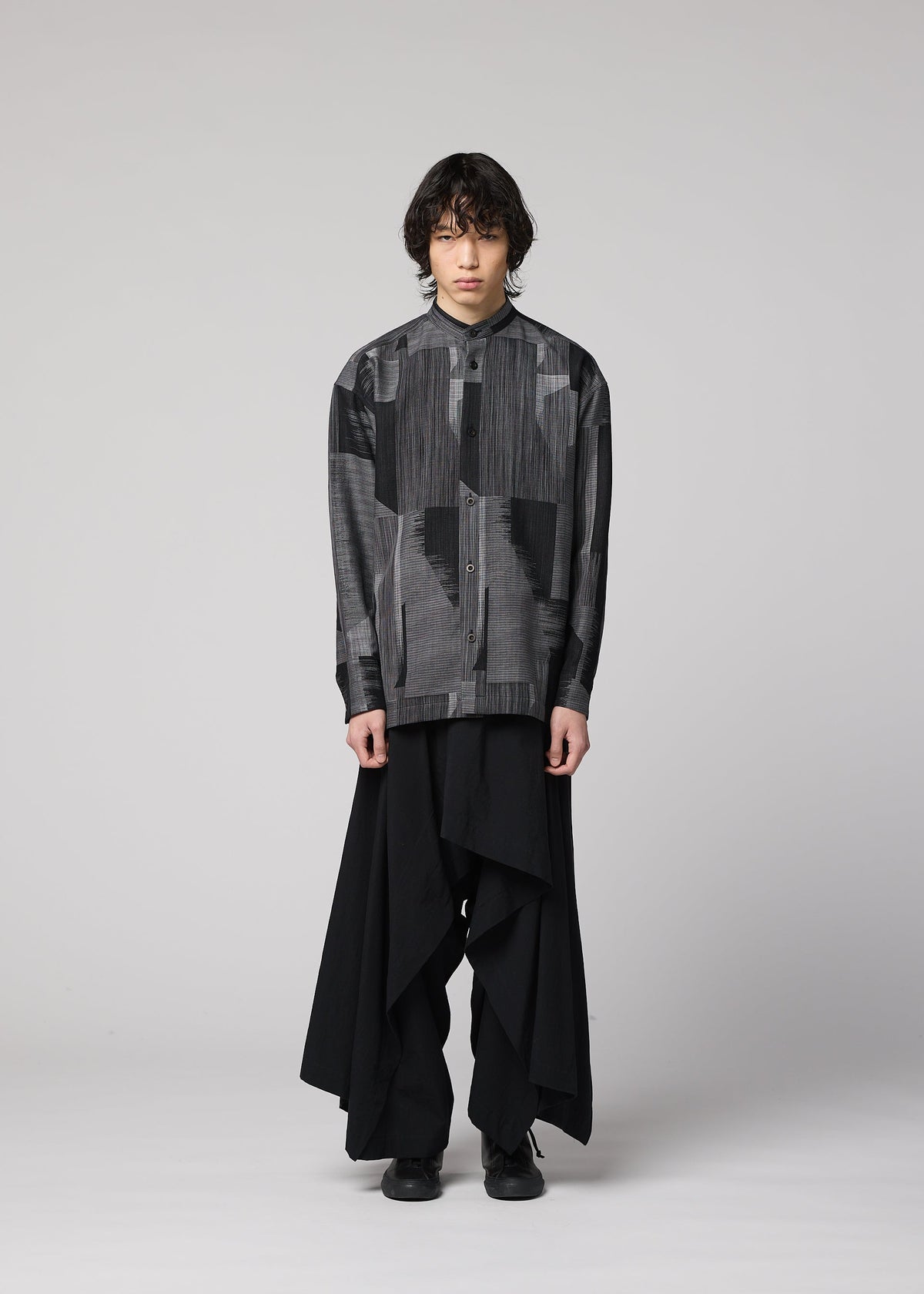 A person stands against a plain background wearing the IM MEN KASURI shirt with a standing collar, paired with wide, layered black pants. Their hair is dark and wavy, and their expression is neutral. view 5 of 8