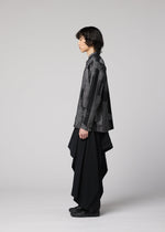 A person stands in profile against a plain background, wearing the KASURI shirt by IM MEN, featuring a black-and-gray pattern with a standing collar, paired with long, draped black pants and black shoes.