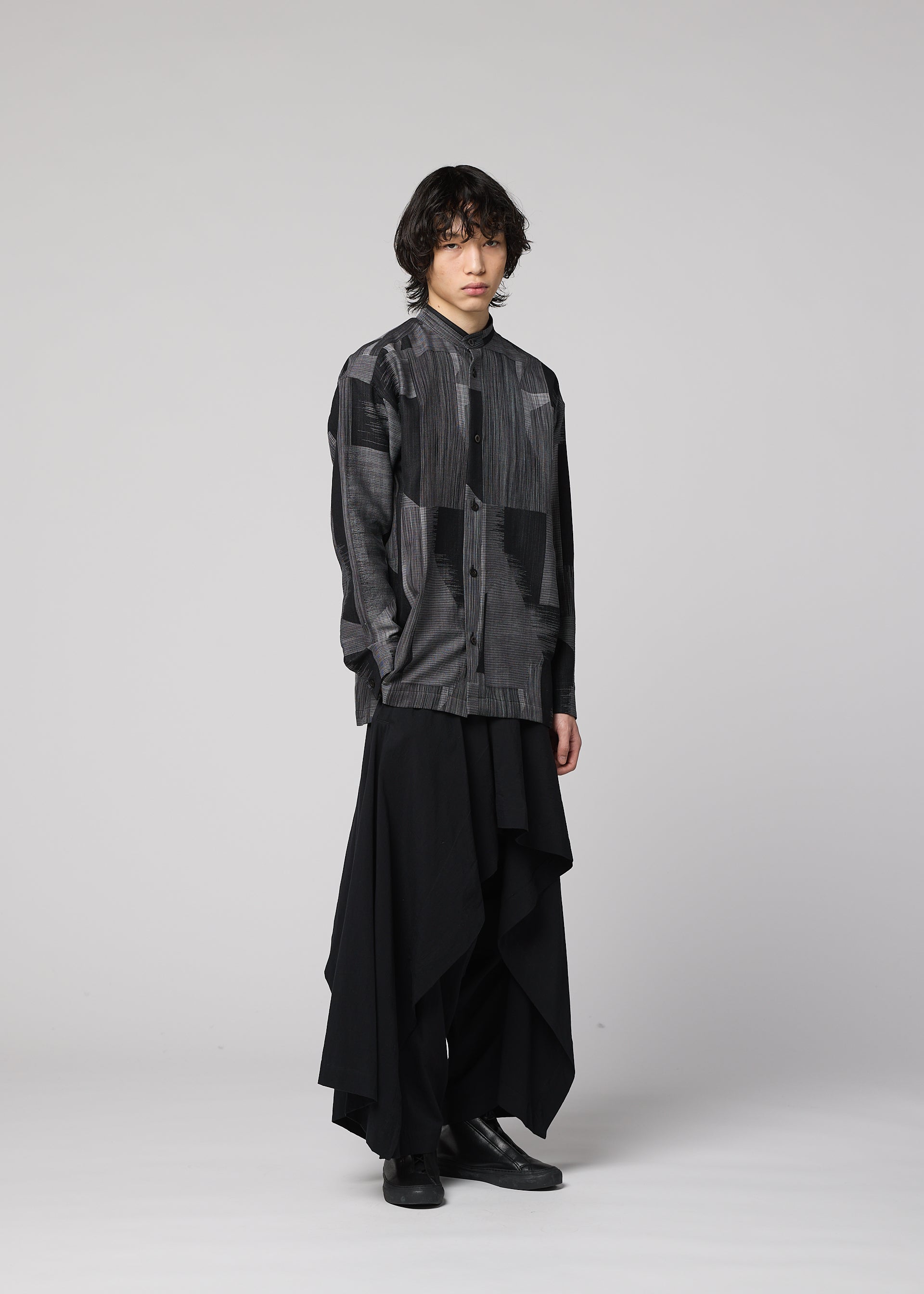 Zoomed view A person with wavy hair wears the IM MEN KASURI shirt in black and gray, featuring intricate Kasuri weaving, paired with long, layered black skirt-like pants, standing against a plain light gray background.