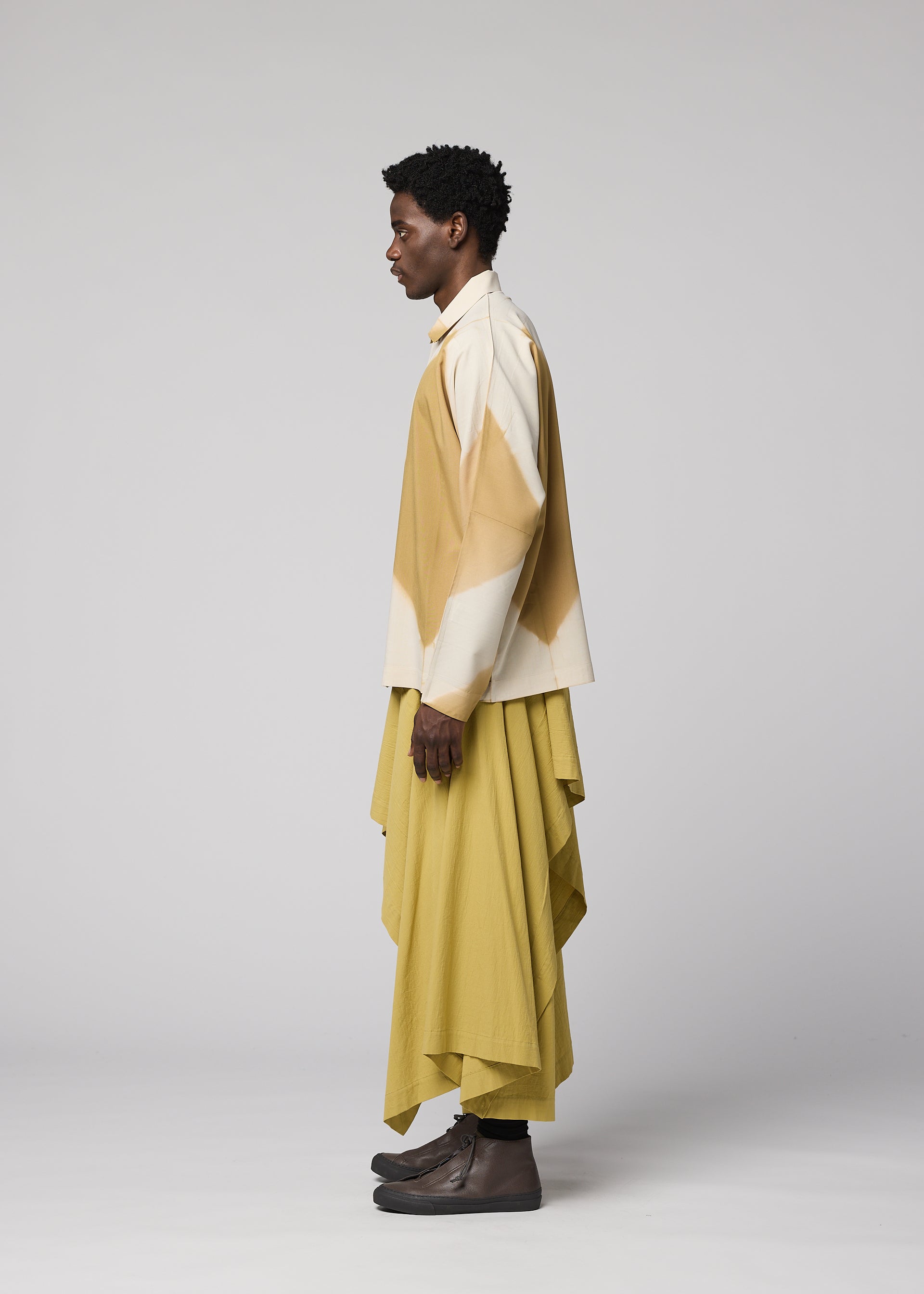 Zoomed view A person stands in profile wearing the ITAJIME SHIRT from IM MEN—a beige and white long-sleeve top showcasing a subtle geometric pattern created using traditional dyeing techniques—paired with a loose, layered yellow skirt and dark slip-on shoes, set against a simple light gray background.