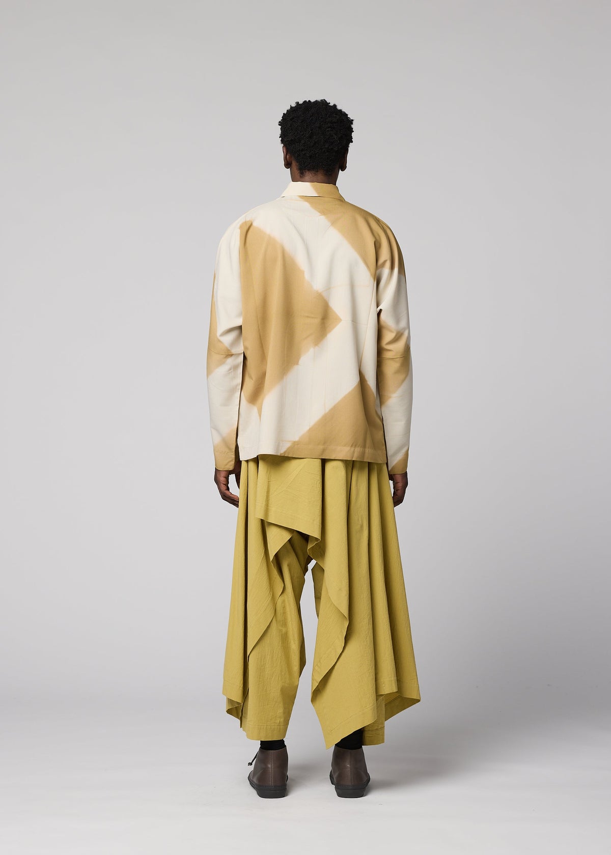 A person stands facing away against a plain light gray background, wearing the IM MEN ITAJIME SHIRT—a patterned beige and white jacket inspired by traditional dyeing techniques—and loose mustard-yellow pants with draped, asymmetrical layers. view 8 of 9