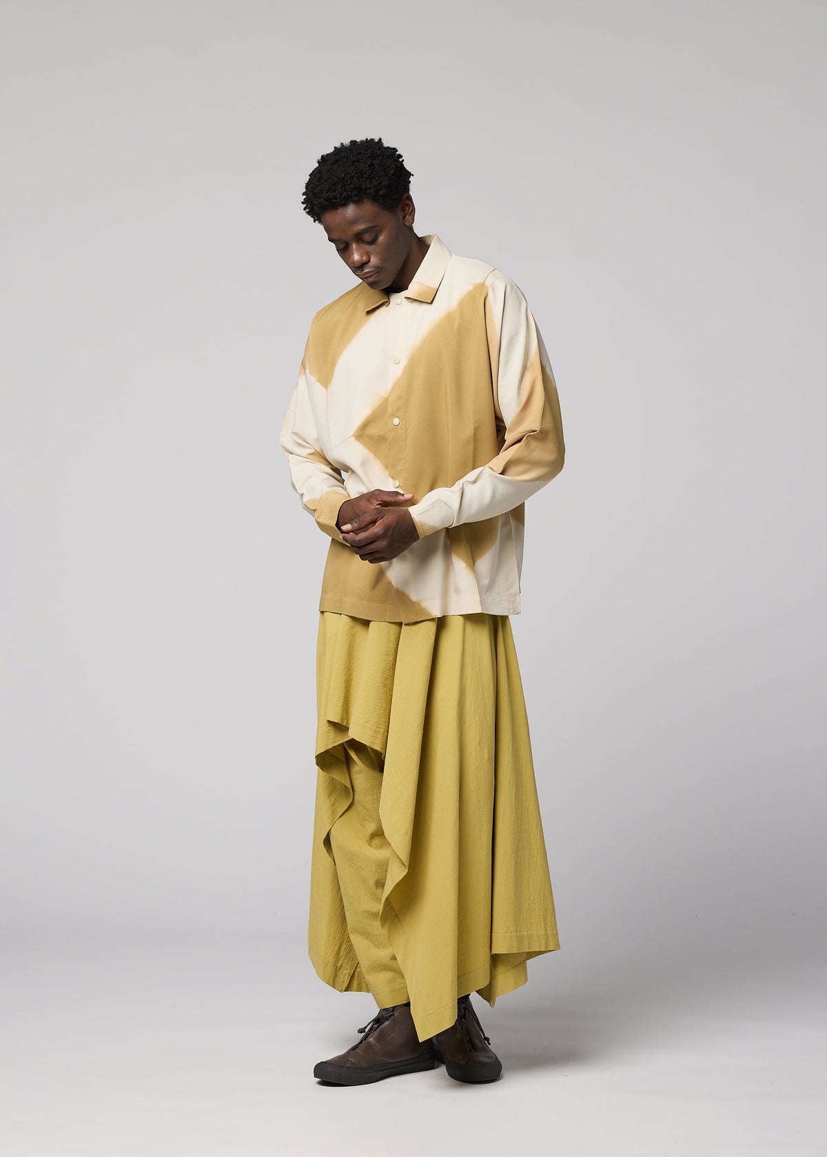 A person stands in a studio wearing the IM MEN ITAJIME SHIRT in beige and cream, paired with a long, flowing yellow skirt and dark shoes. They look down with hands clasped against a plain light gray background. view 9 of 9