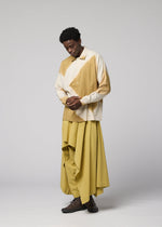 A person stands in a studio wearing the IM MEN ITAJIME SHIRT in beige and cream, paired with a long, flowing yellow skirt and dark shoes. They look down with hands clasped against a plain light gray background.