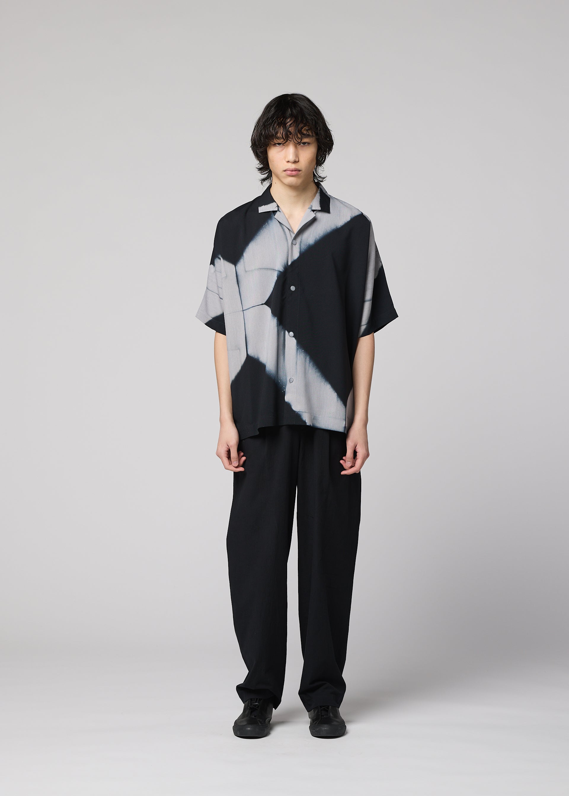 Zoomed view A person with medium-length dark hair stands facing forward, dressed in the IM MEN ITAJIME SHIRT—a black and gray short-sleeve button-up featuring subtle geometric patterns inspired by the traditional Itajime Shibori dyeing technique—paired with loose black pants and black shoes against a light gray background.