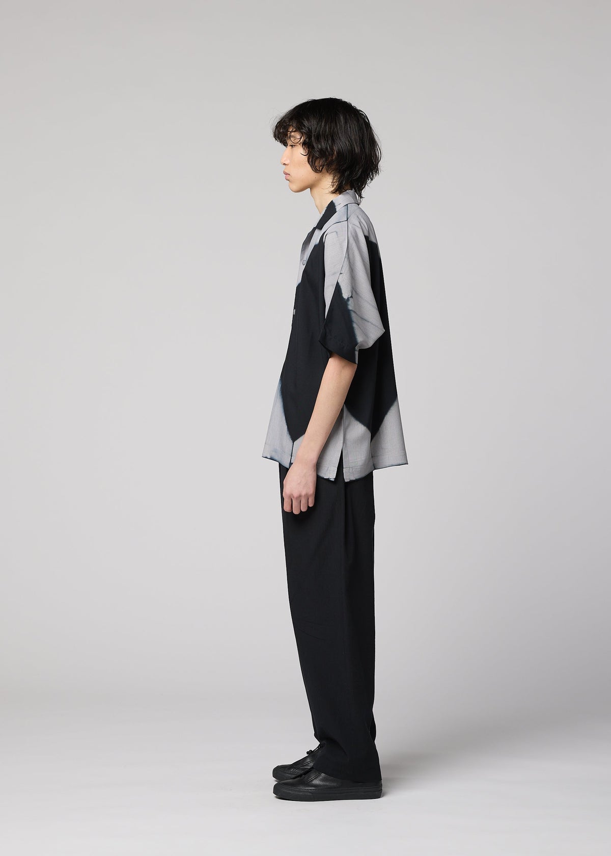 A person with medium-length dark hair stands in profile against a plain light gray background, wearing the IM MEN ITAJIME SHIRT—a short-sleeved button-up featuring subtle geometric patterns inspired by traditional Itajime Shibori dyeing—paired with black pants and black shoes. view 8 of 10