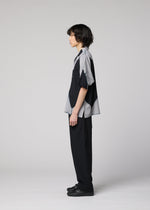 A person with medium-length dark hair stands in profile against a plain light gray background, wearing the IM MEN ITAJIME SHIRT—a short-sleeved button-up featuring subtle geometric patterns inspired by traditional Itajime Shibori dyeing—paired with black pants and black shoes.