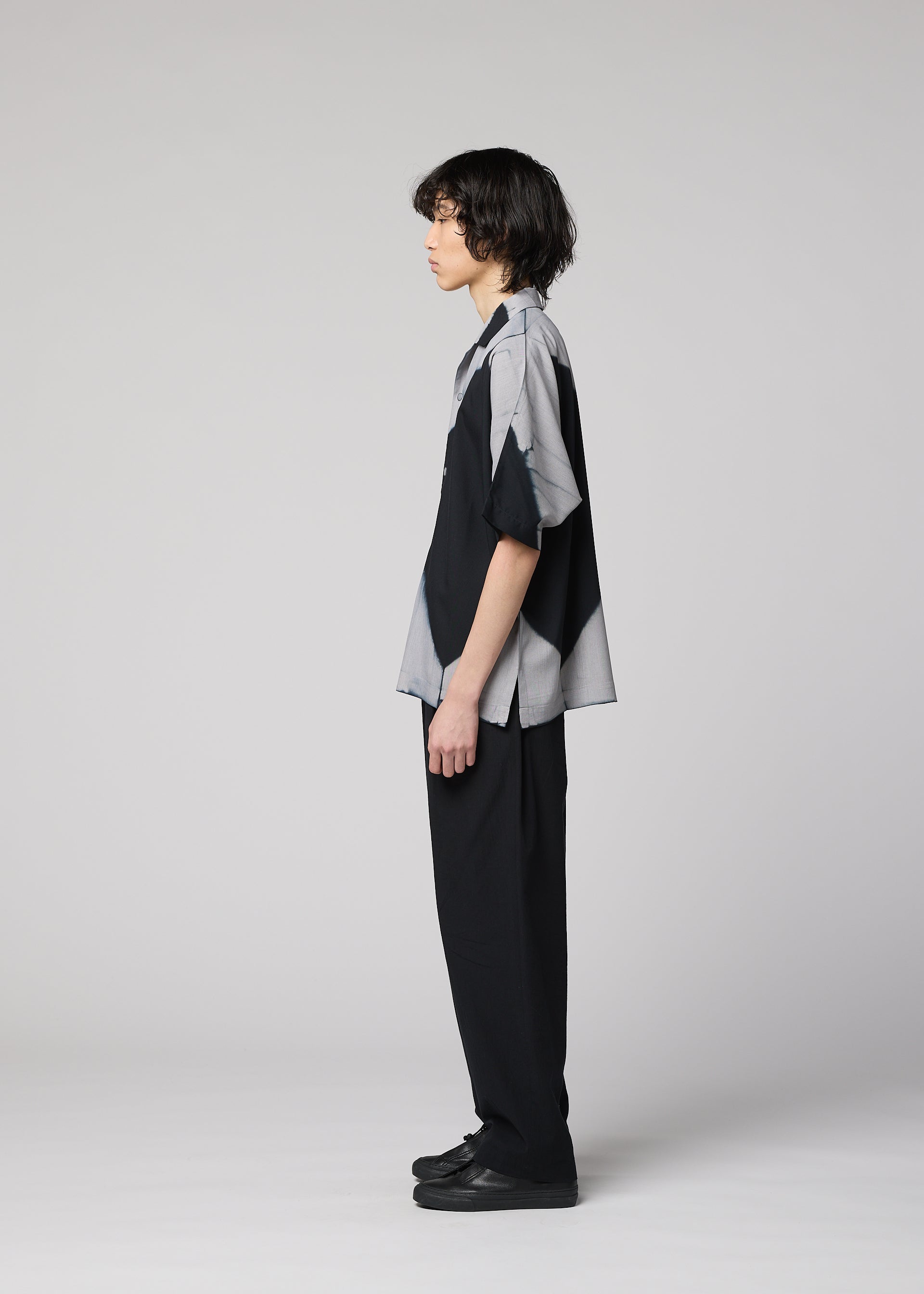 Zoomed view A person with medium-length dark hair stands in profile against a plain light gray background, wearing the IM MEN ITAJIME SHIRT—a short-sleeved button-up featuring subtle geometric patterns inspired by traditional Itajime Shibori dyeing—paired with black pants and black shoes.