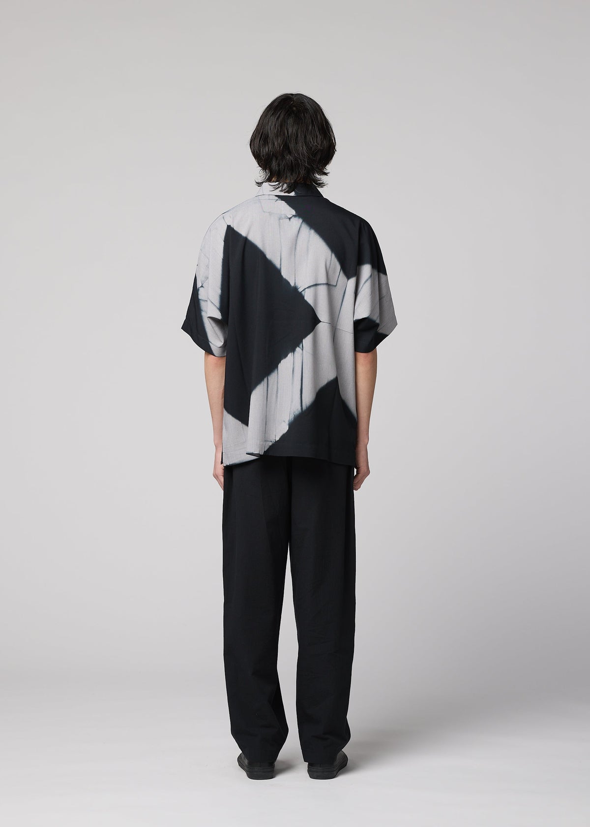 A person with medium-length dark hair stands facing away, wearing the IM MEN ITAJIME SHIRT—a loose, short-sleeved black and white shirt featuring geometric patterns inspired by traditional Itajime Shibori—paired with black pants, against a plain light gray background. view 9 of 10