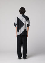 A person with medium-length dark hair stands facing away, wearing the IM MEN ITAJIME SHIRT—a loose, short-sleeved black and white shirt featuring geometric patterns inspired by traditional Itajime Shibori—paired with black pants, against a plain light gray background.