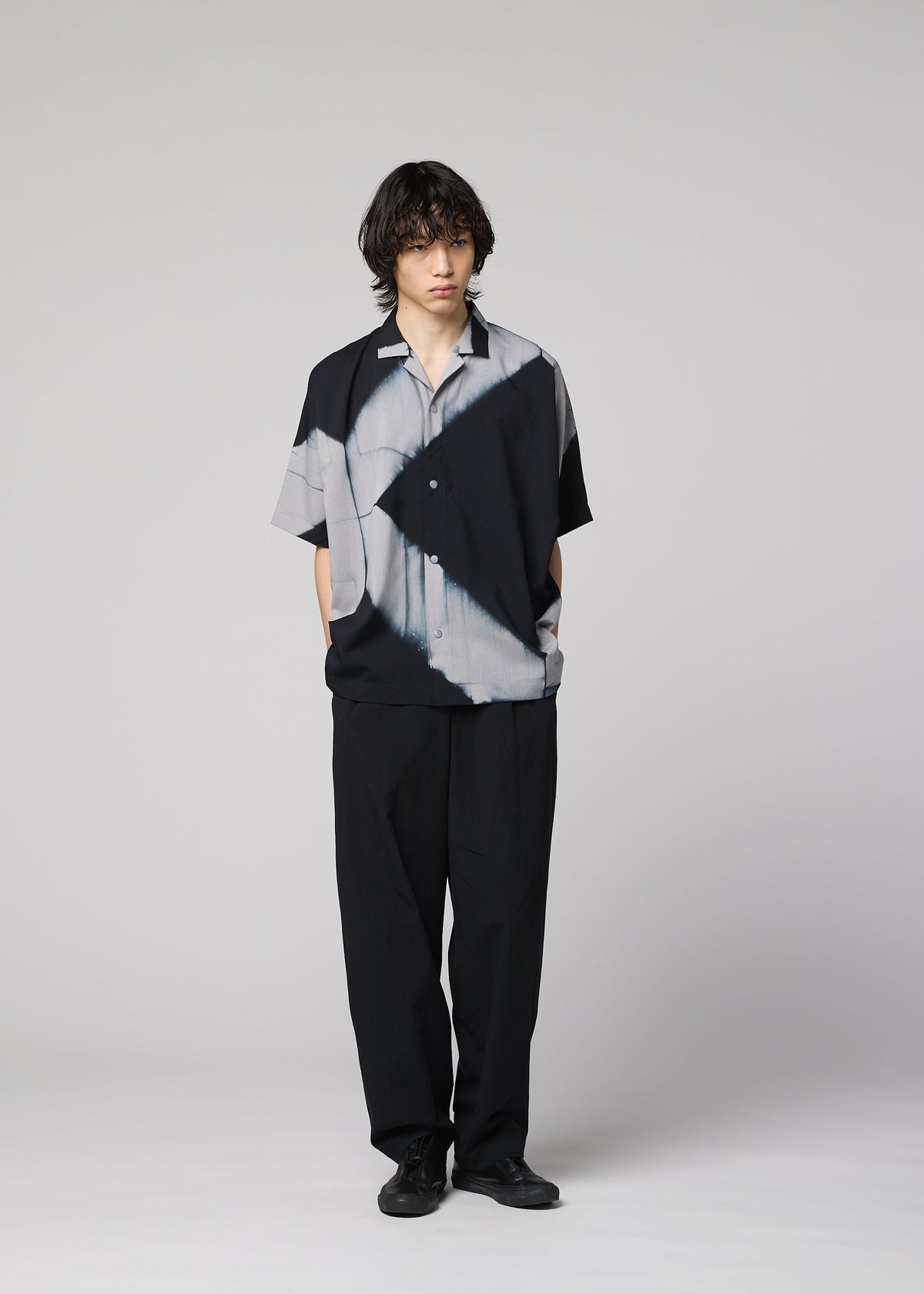 Against a plain background, a person with medium-length dark hair and a neutral expression wears the ITAJIME SHIRT by IM MEN—an eye-catching black and gray short-sleeve top featuring geometric patterns inspired by Itajime Shibori, a traditional dyeing technique. The shirt is paired with loose black pants and black shoes. view 10 of 10