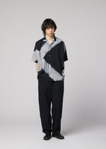 Against a plain background, a person with medium-length dark hair and a neutral expression wears the ITAJIME SHIRT by IM MEN—an eye-catching black and gray short-sleeve top featuring geometric patterns inspired by Itajime Shibori, a traditional dyeing technique. The shirt is paired with loose black pants and black shoes.