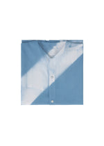 The ITAJIME SHIRT by IM MEN is a folded light blue shirt showcasing geometric patterns inspired by the traditional Itajime Shibori dyeing technique. It features visible snap buttons and a white diagonal shadow, and is displayed against a white background.