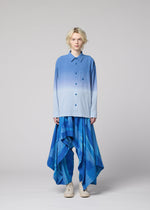 A person with light hair wears the IM MEN DRIP SHIRT, a blue gradient dye button-up, paired with voluminous blue patterned pants. They stand against a plain gray background, facing the camera with a neutral expression.