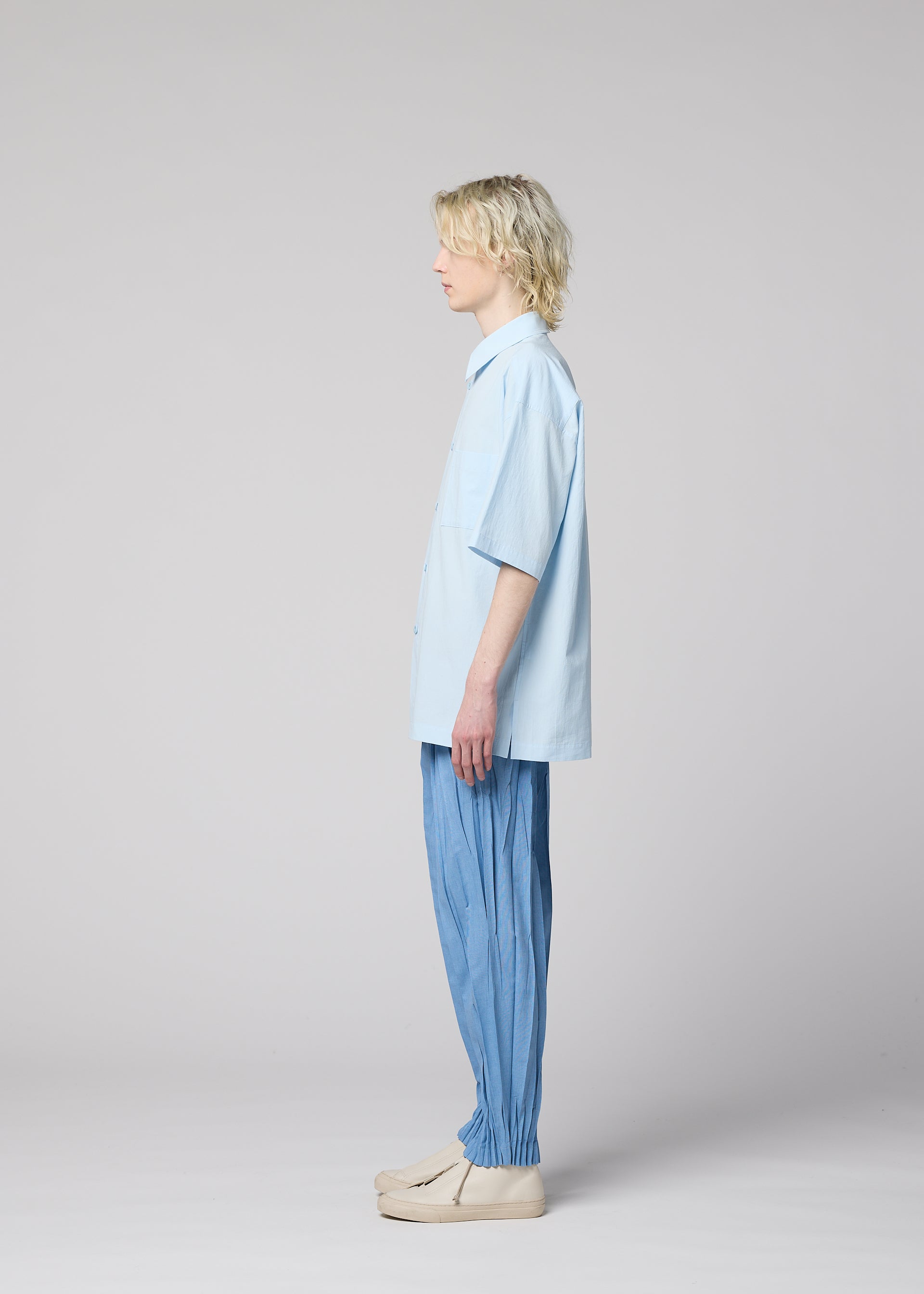 Zoomed view A person with light hair stands in profile against a plain background, wearing the IM MEN WHITE SHIRT—a light blue, regular-collar, short-sleeved shirt crafted from cotton linen ramie—paired with loose blue pants gathered at the ankles and beige shoes.