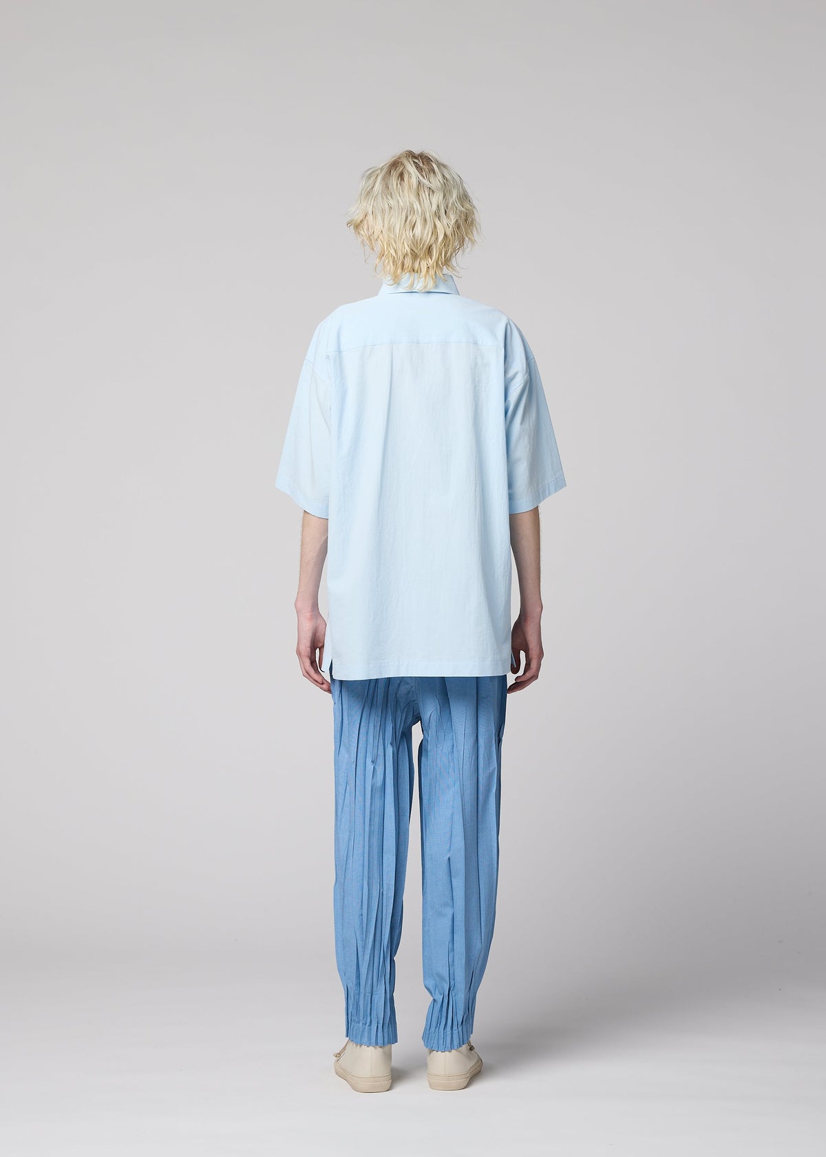 A person with curly blond hair stands facing away, wearing the IM MEN WHITE SHIRT—a loose, short-sleeved cotton linen ramie top—paired with pleated blue pants gathered at the ankles and beige shoes, set against a plain light gray background. view 8 of 9
